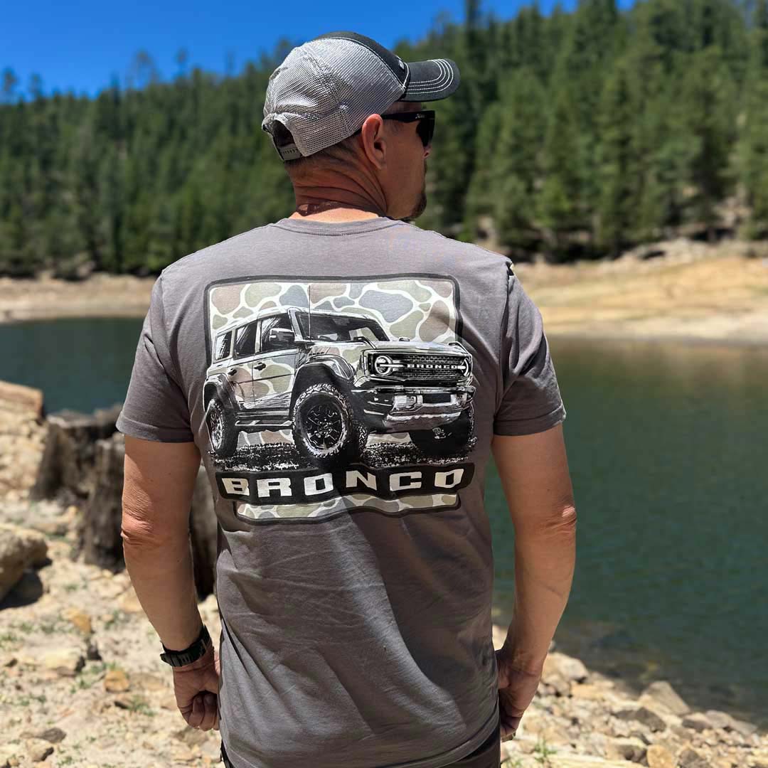 A man wearing a gray t-shirt with a large back graphic of a Ford Bronco SUV, photographed outdoors by a lake with a forest in the background.