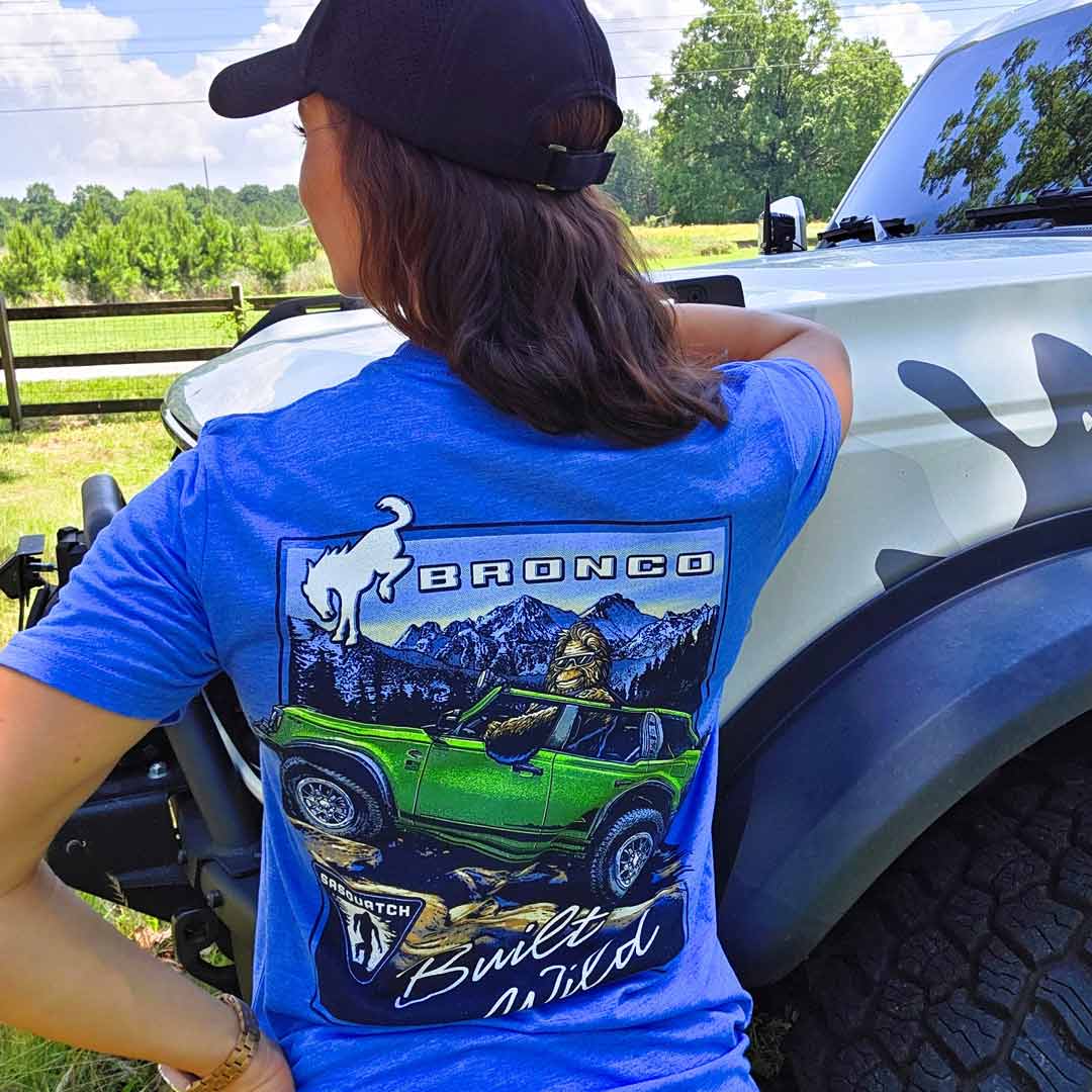 Woman wearing blue Bronco Sasquatch T-Shirt in front of a grassy environment and a Silver Bronco