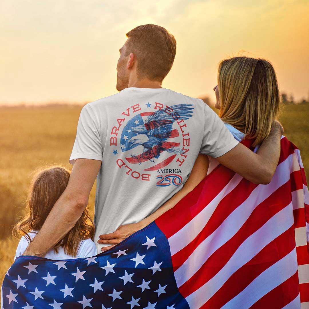 Family of three standing in a field with an American flag draped over them, featuring an America 250 t-shirt with an eagle design.