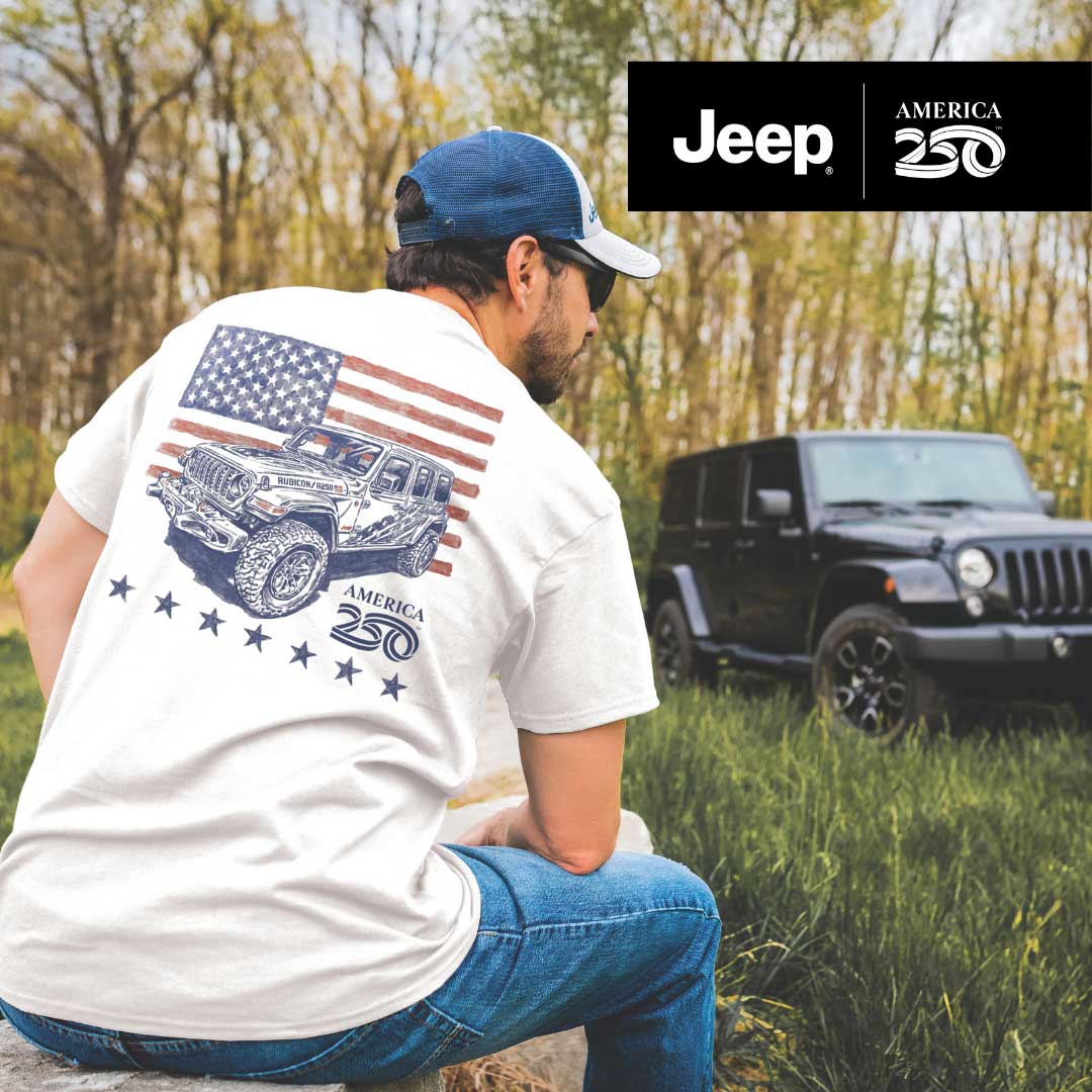 Man wearing a white t-shirt with Jeep graphics and an American flag, sitting in a grassy field with a Jeep in the background.