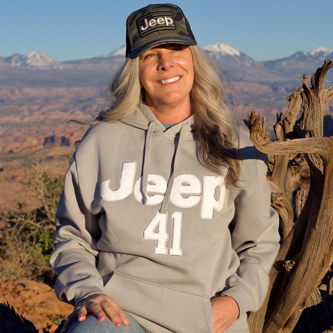 A woman wearing a gray hoodie with 'Jeep 41' printed on the front, blue jeans, and a camouflage cap with a Jeep logo, posing outdoors in a desert landscape with rock formations and mountains in the background.