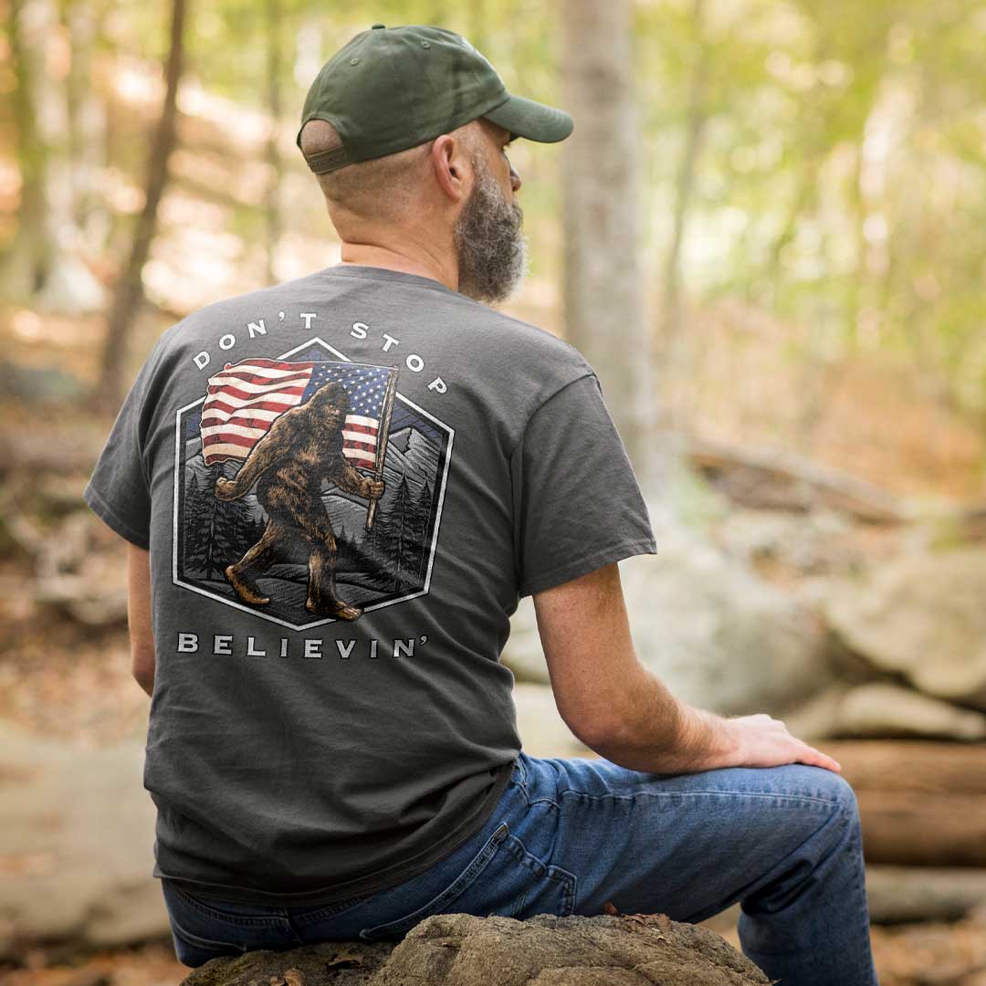 man sitting in the woods wearing a gray t-shirt with sasquatch and American flag