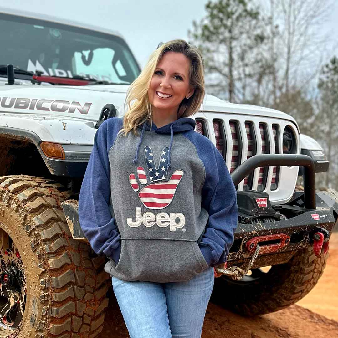 Woman in front of a jeep wearing a jeep themed hoodie that has a hand waving with the American flag overlayed on it