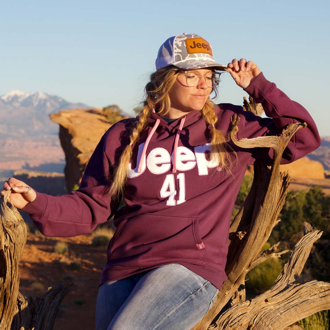 A woman wearing a maroon hoodie with 'Jeep 41' printed on the front, blue jeans, and a camouflage cap with a Jeep logo, posing outdoors in a desert landscape with rock formations and mountains in the background.